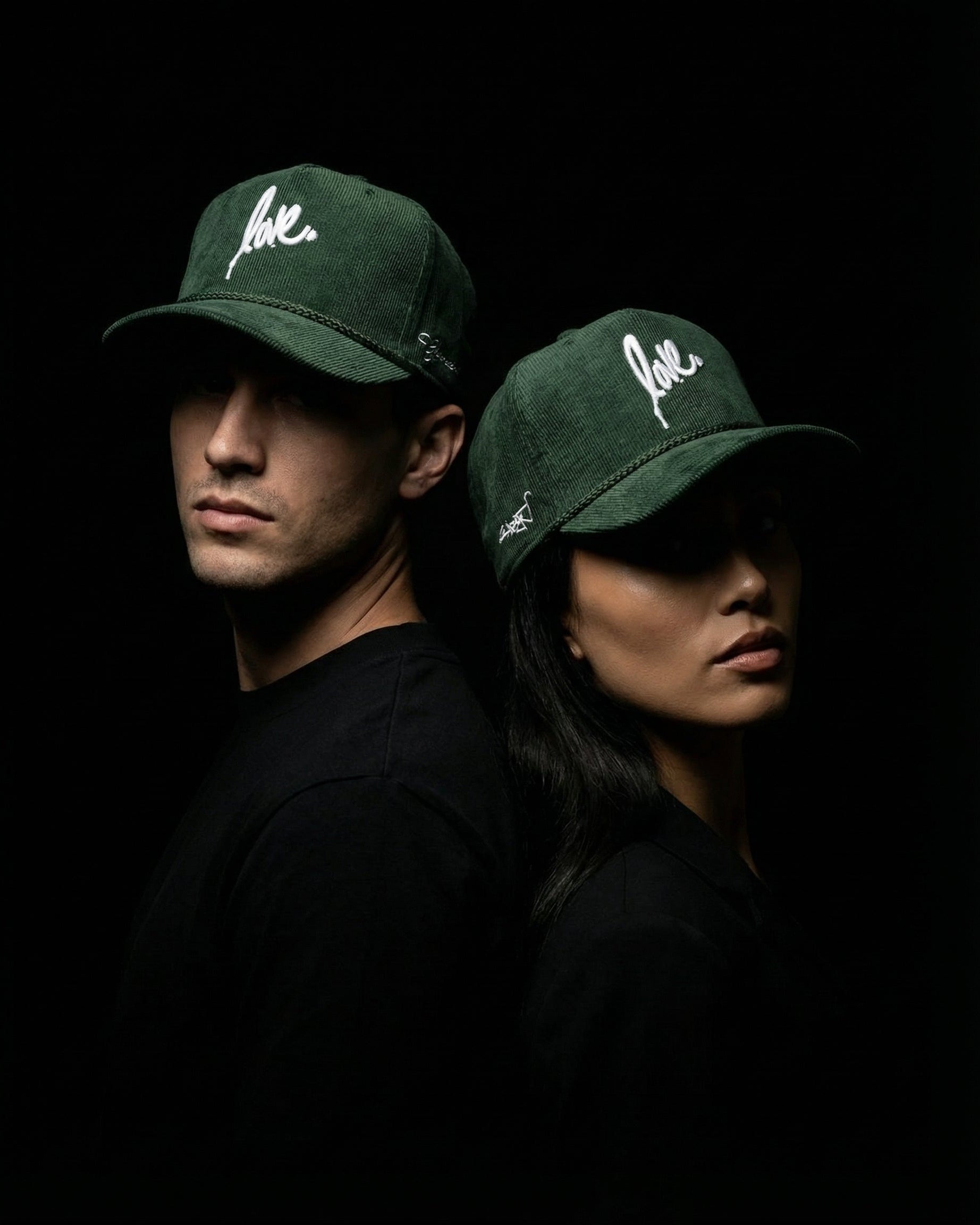 Two people wearing green corduroy ruben rojas Live Through Love caps with 'love' printed on them against a black background