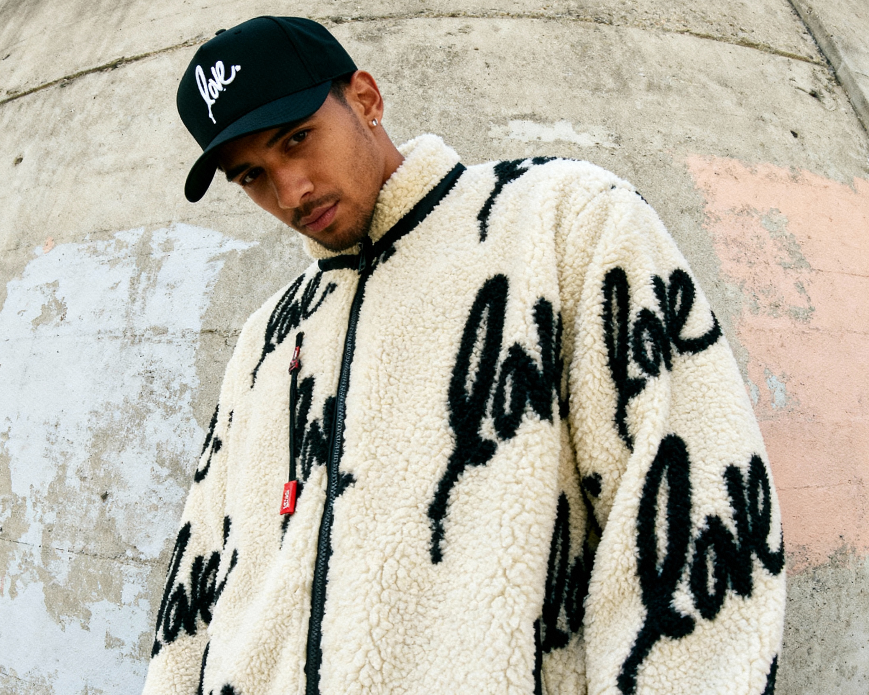 Person wearing a white jacket with 'love' text, black cap, and pants against a concrete wall.