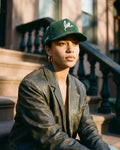 Person wearing a green corduroy love cap and leather jacket sitting on steps outdoors