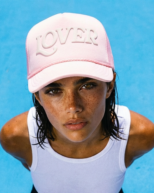 Person wearing a white tank top and pink ruben rojas cap with 'LOVER' text, standing in clear blue water.