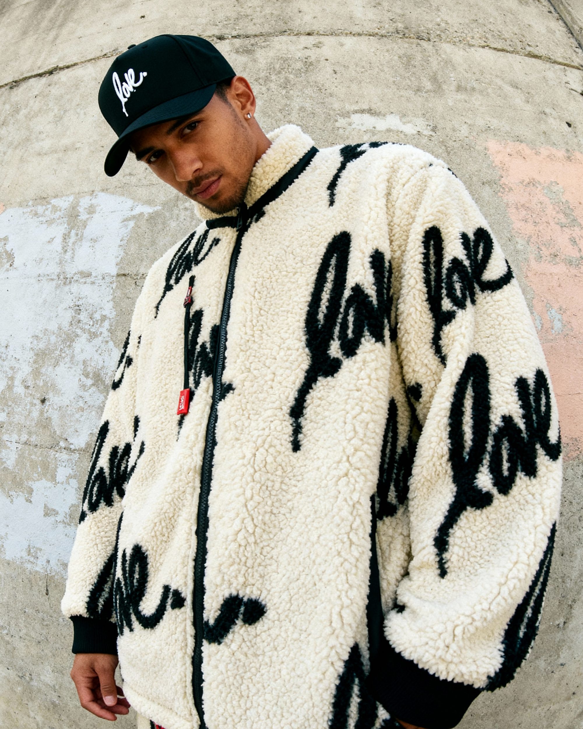 Person wearing a white jacket with 'love' text, black cap, and pants against a concrete wall.