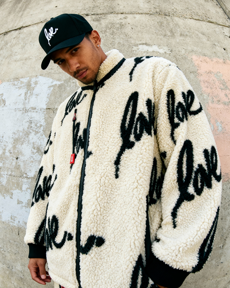 Person wearing a white jacket with 'love' text, black cap, and pants against a concrete wall.